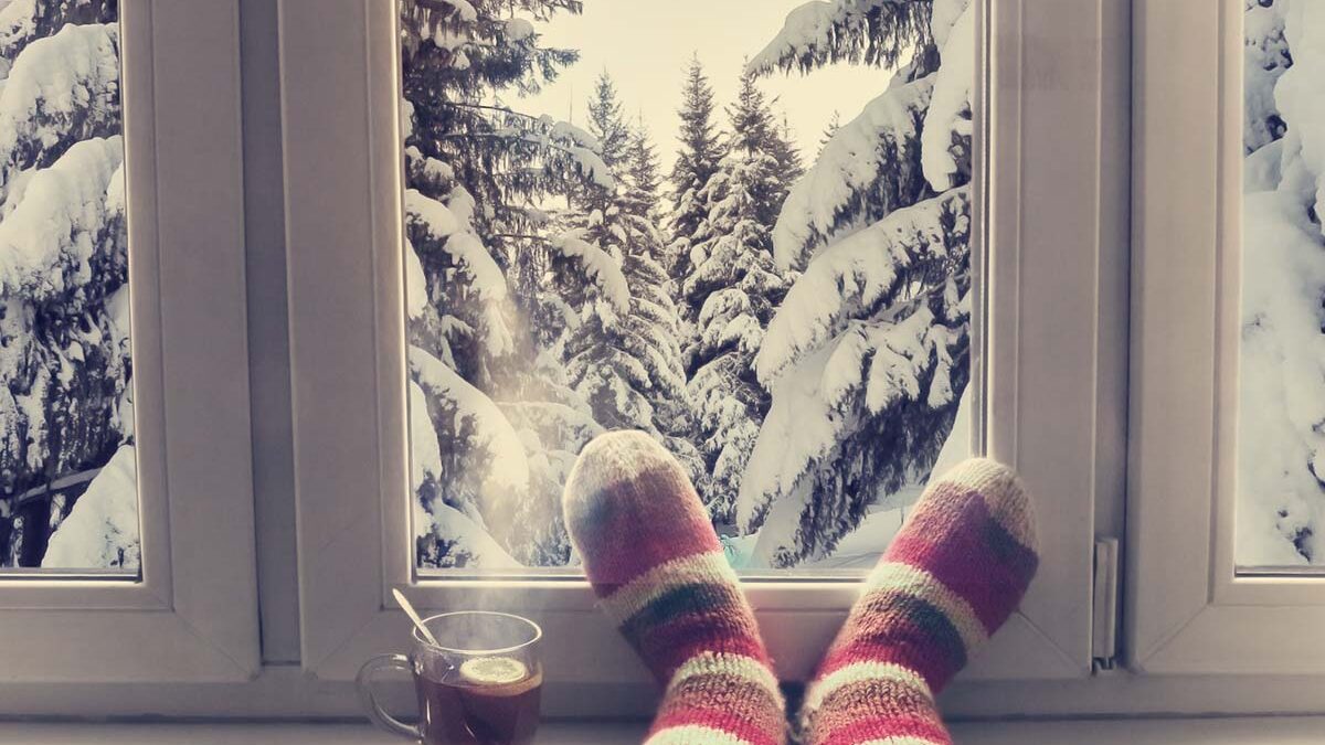 a set of striped socks sits in front of a window showing a wintery scene of snow on pine trees after the person learns how to winterize windows