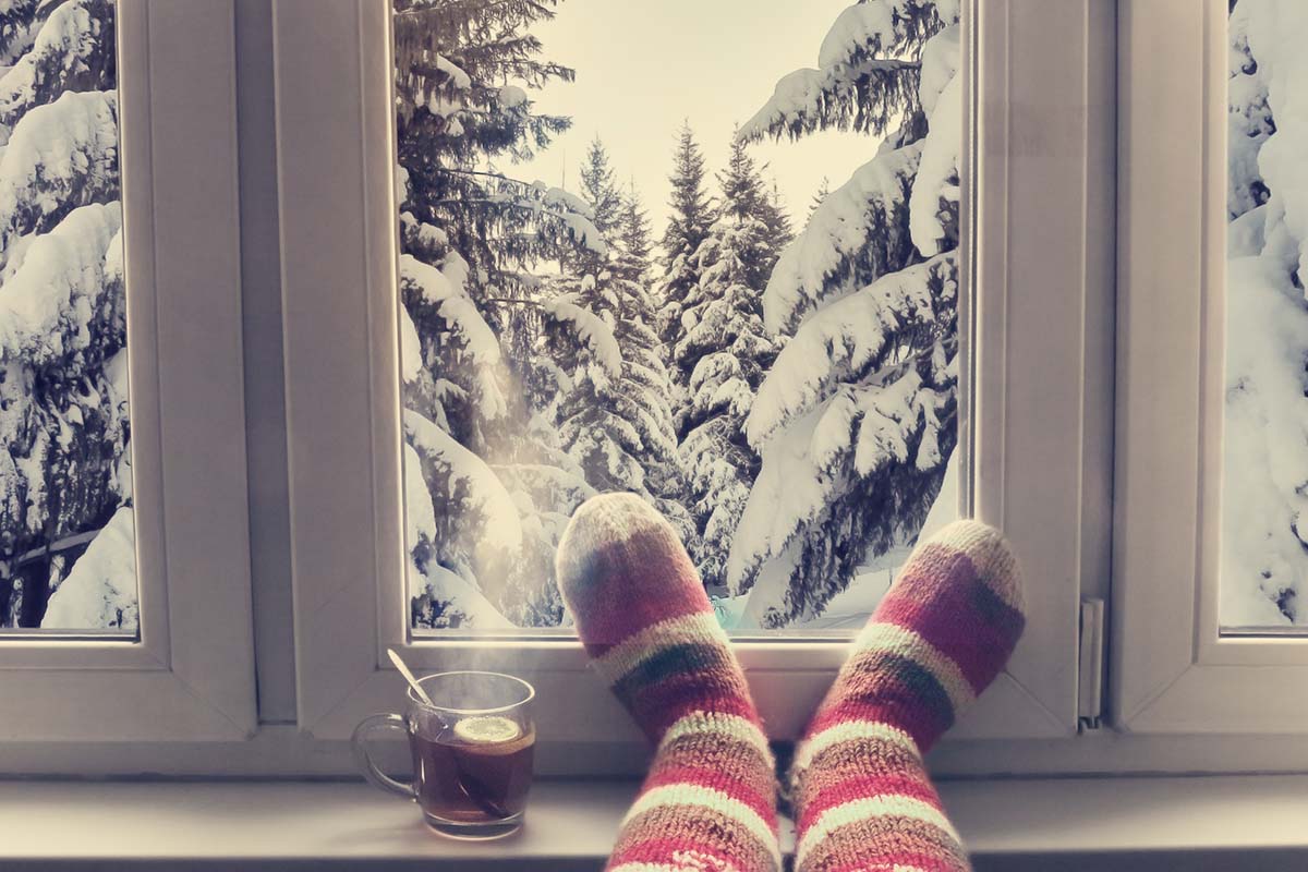 a set of striped socks sits in front of a window showing a wintery scene of snow on pine trees after the person learns how to winterize windows