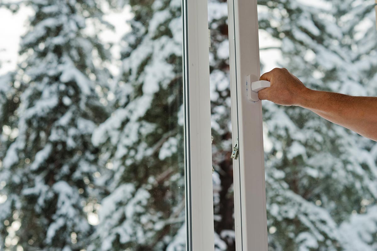 Should I Prepare My Windows for Winter_ a picture of someone opening a window to a snowy set of pine trees after they prepare windows for winter