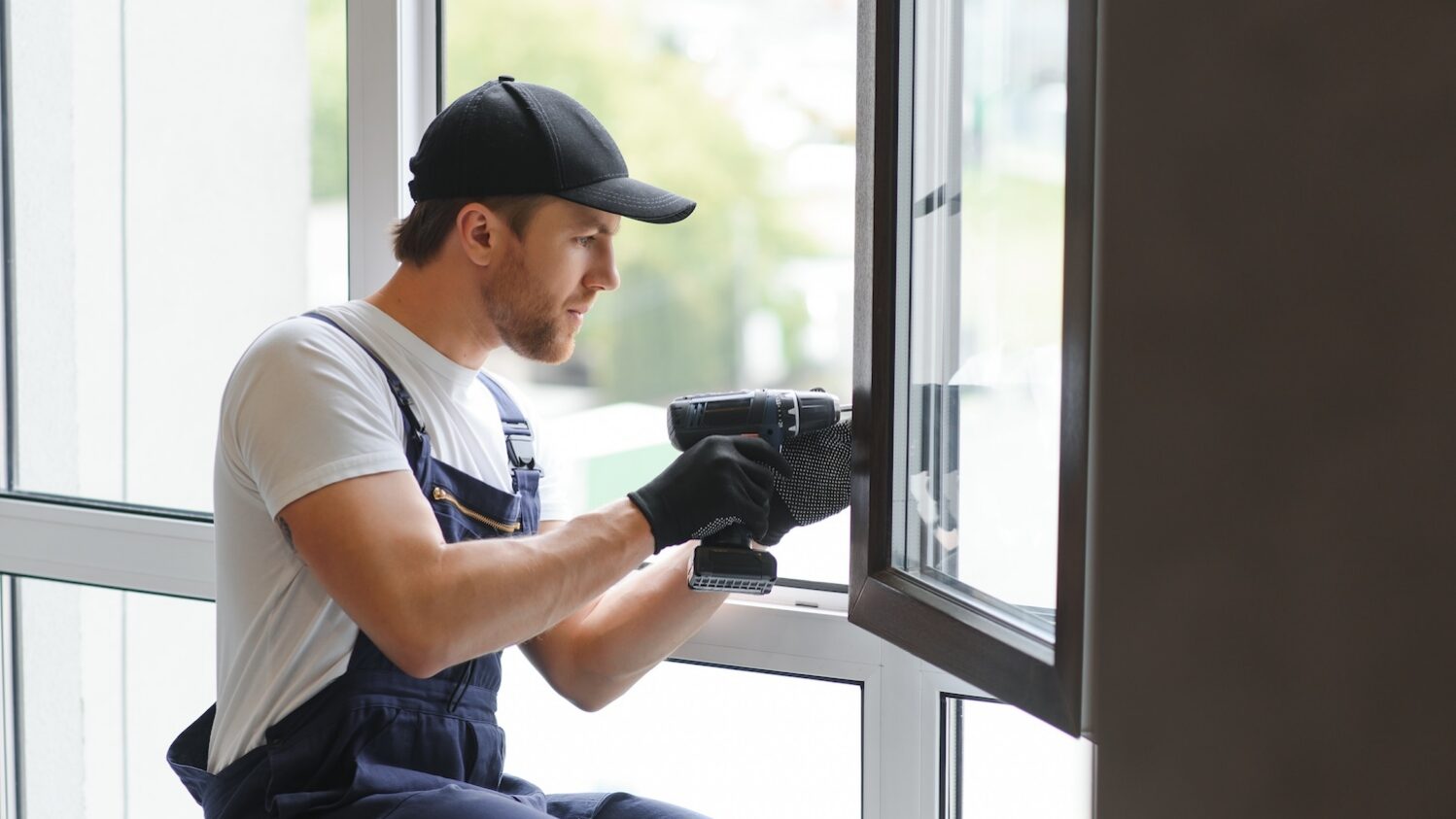Worker wearing overalls, gloves, and a cap using a power drill to install or repair a window frame indoors, concentrating on the task in a bright, modern space.