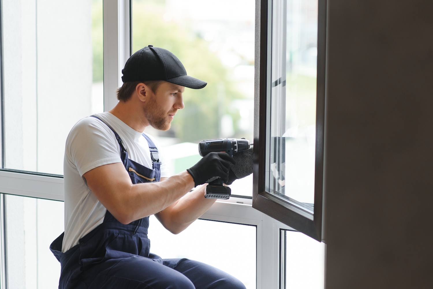 Construction worker installing window in house. Worker wearing overalls, gloves, and a cap using a power drill to install or repair a window frame indoors, concentrating on the task in a bright, modern space.