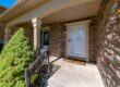 Front entrance of a suburban house with a white door, stone exterior walls, and a small covered porch featuring a railing, a welcome mat, and greenery along the walkway.