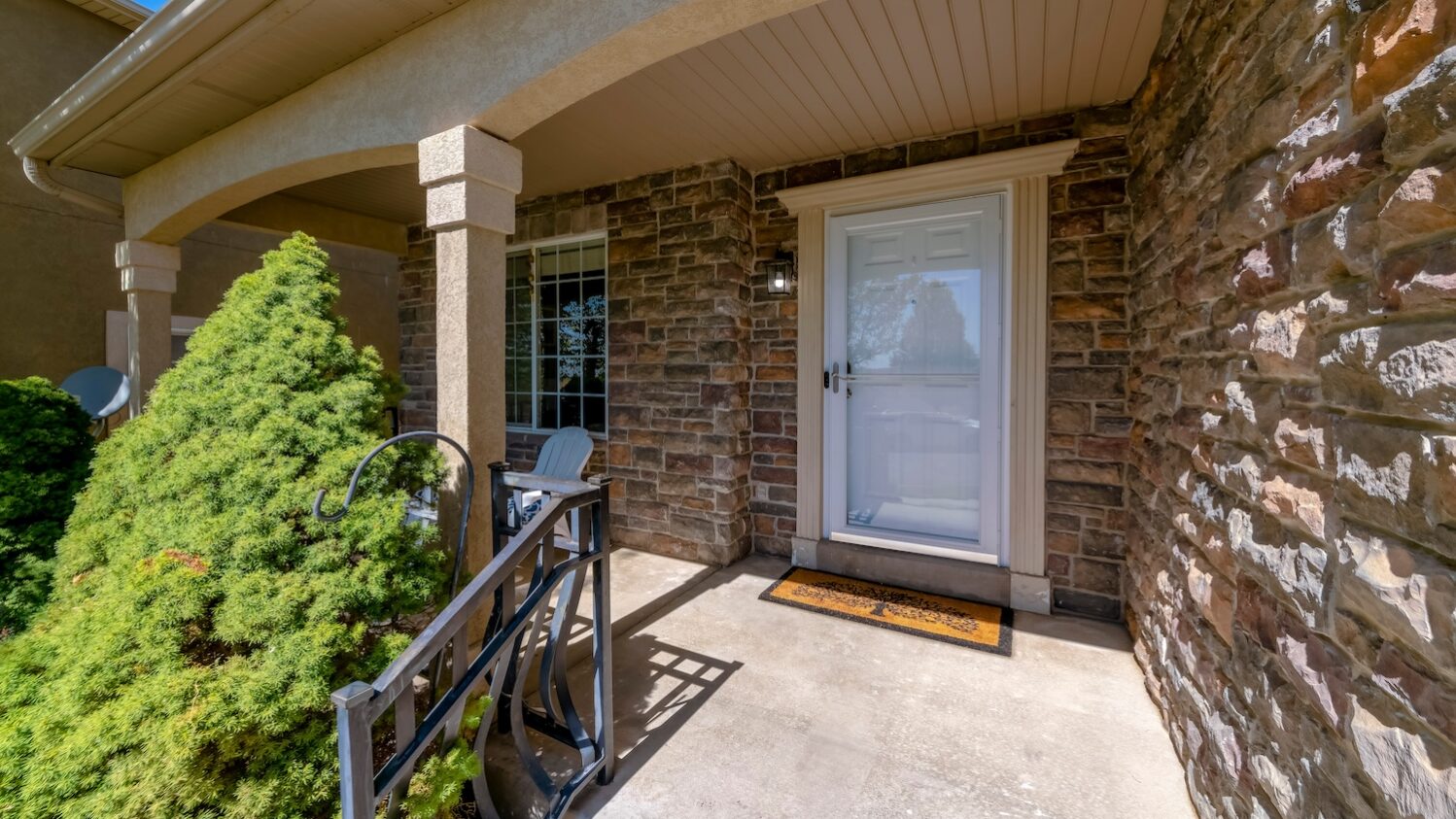 Front entrance of a suburban house with a white door, stone exterior walls, and a small covered porch featuring a railing, a welcome mat, and greenery along the walkway.