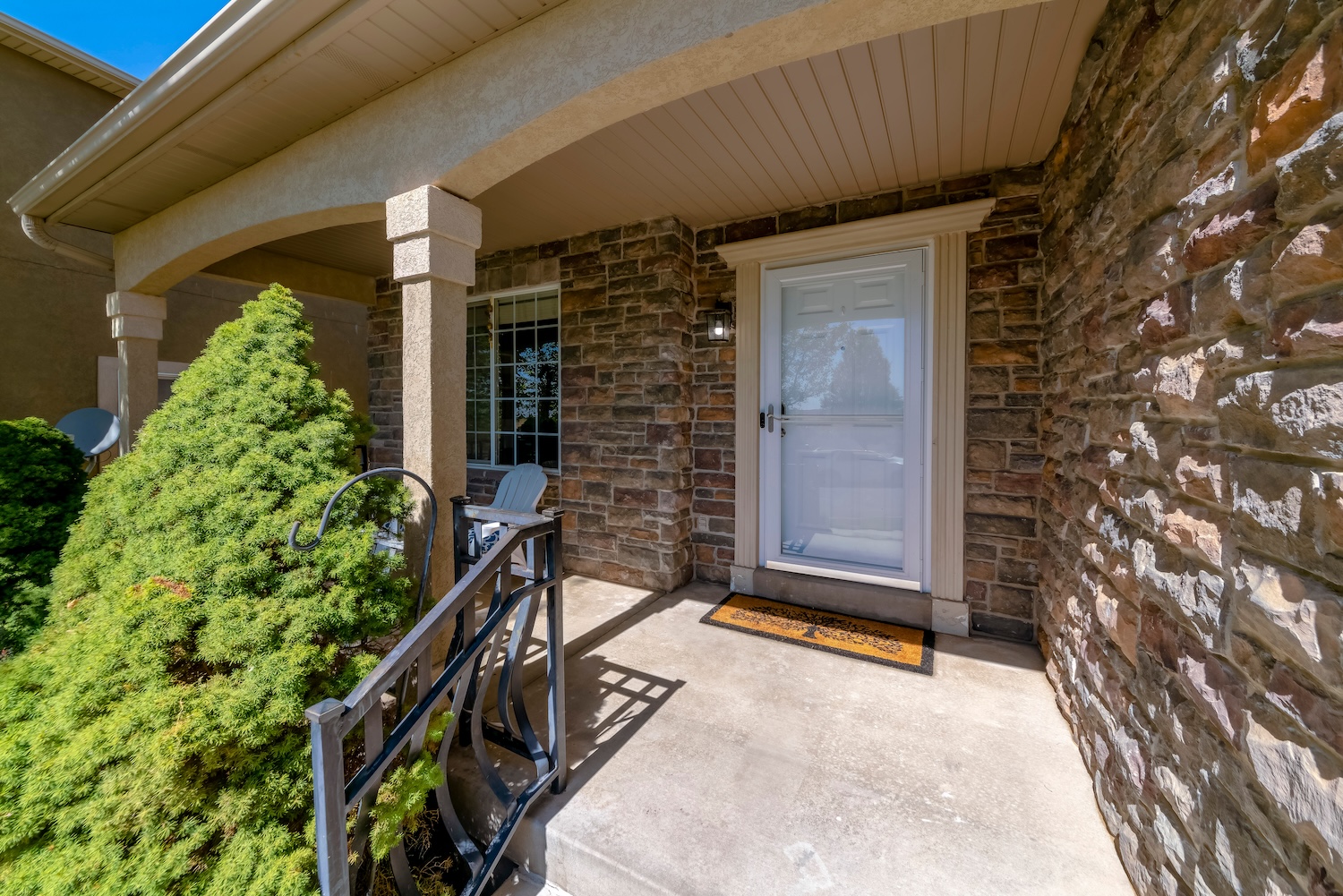 Front entrance of a suburban house with a white door, stone exterior walls, and a small covered porch featuring a railing, a welcome mat, and greenery along the walkway.