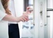 Woman in a white blazer unlocking a modern silver door with a key, focusing on her hands and the sleek metal lock mechanism.