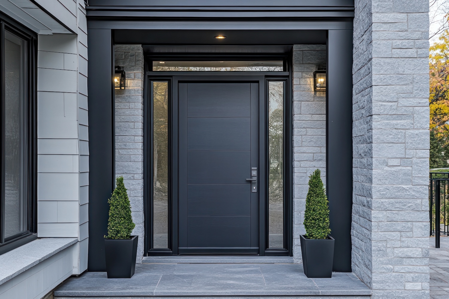 Modern front entrance featuring a sleek black door framed by light gray brick and black trim, with two matching potted topiary plants on either side and wall-mounted lights above.