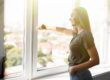 Woman standing by a large bright window, smiling as she opens it to let in fresh air and sunlight, with a soft cityscape visible in the background.