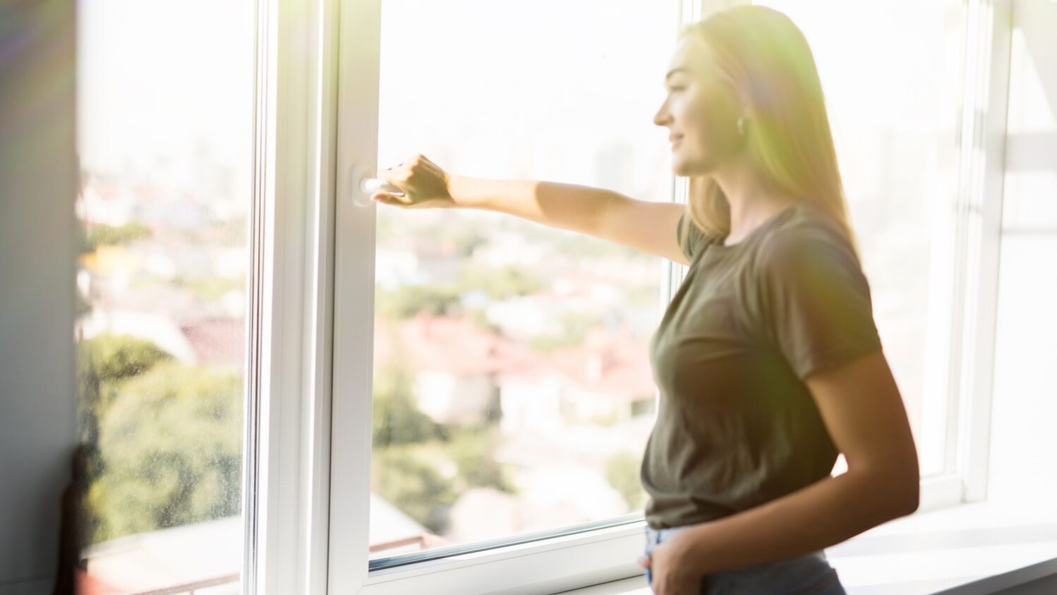 Woman standing by a large bright window, smiling as she opens it to let in fresh air and sunlight, with a soft cityscape visible in the background.
