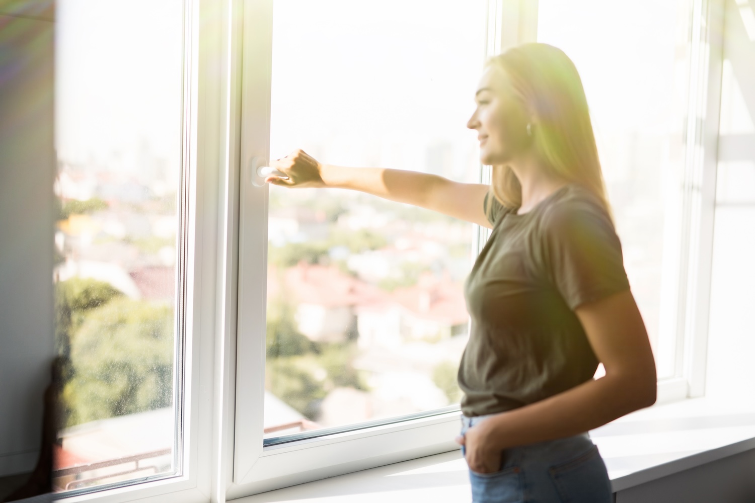 Woman standing by a large bright window, smiling as she opens it to let in fresh air and sunlight, with a soft cityscape visible in the background.