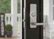Close-up of a modern smart door lock on a black door slightly open, showing a bright porch with a white railing and a “Welcome” sign in the background.