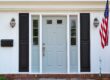 Light blue front entry door with glass sidelights on a white house with black shutters.