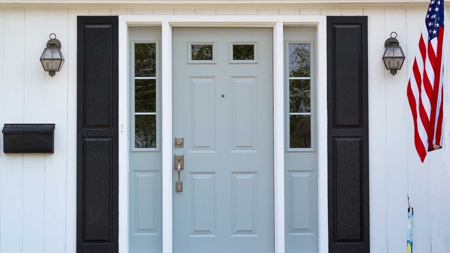 Light blue front entry door with glass sidelights on a white house with black shutters.