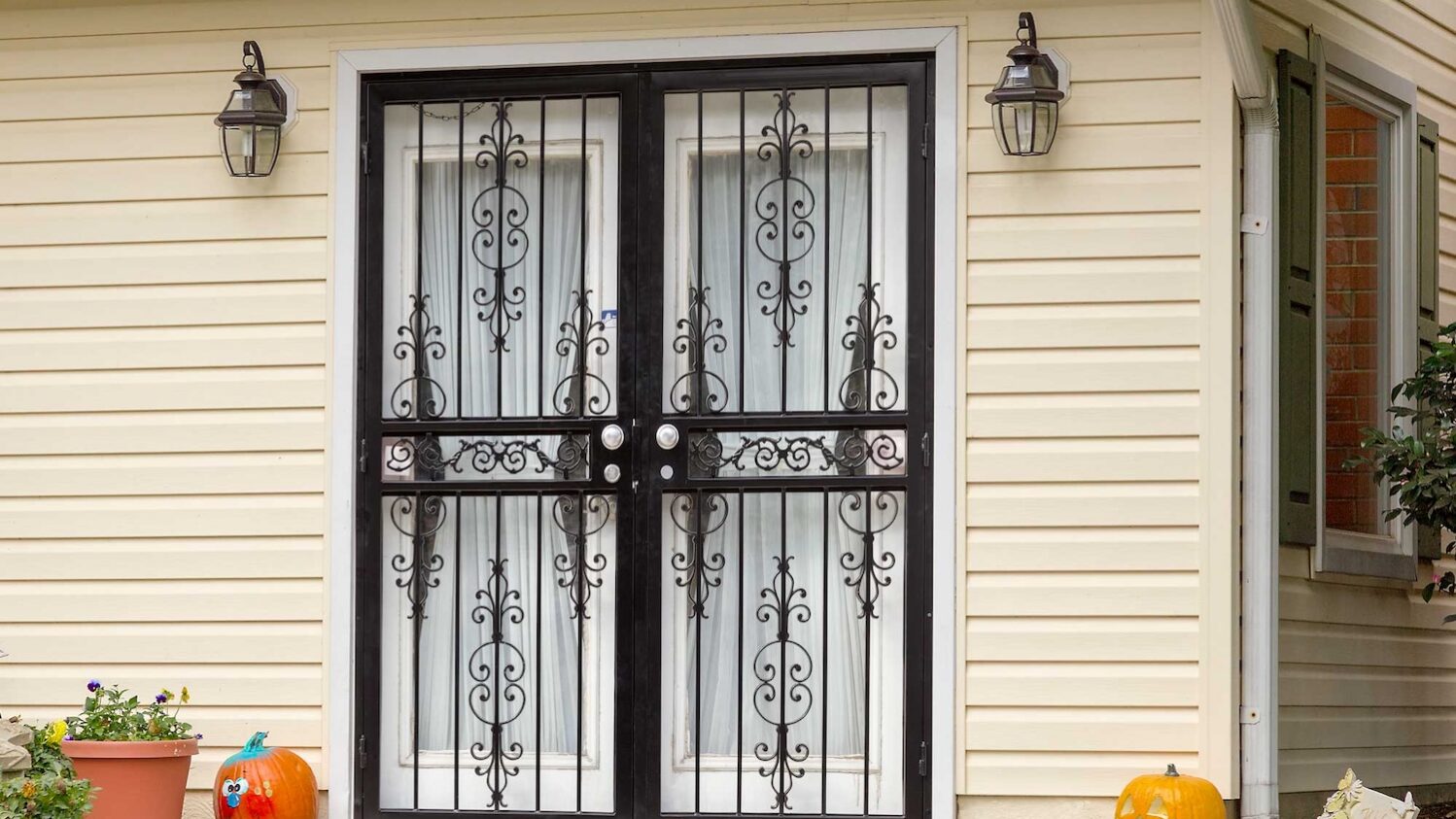 Decorative black wrought iron security doors on a light yellow home with pumpkins outside.
