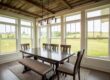 Dining room surrounded by large white-framed windows overlooking a grassy landscape